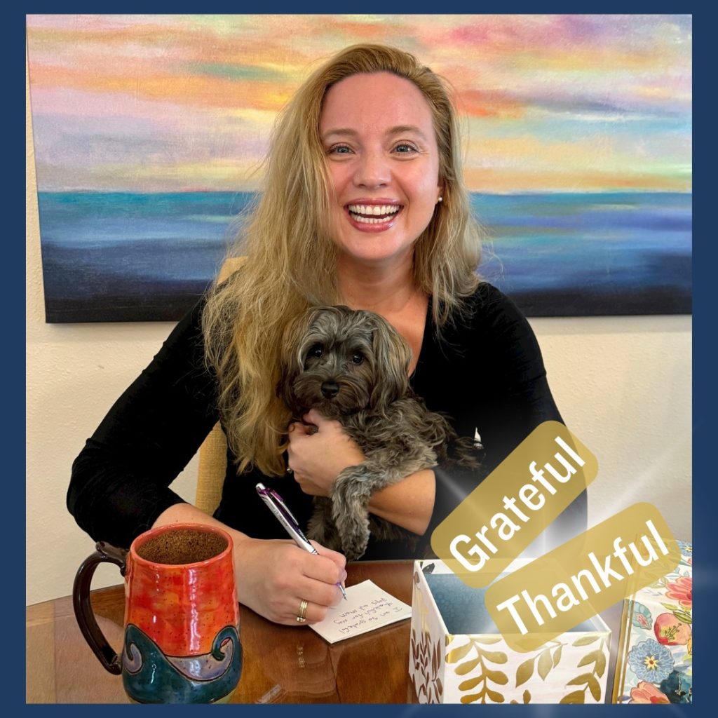 Laura Marie smiling and sitting at a table with her dog Mira in her arms. The words Grateful and Thankful are visible.